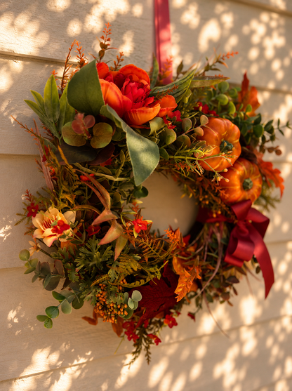 The Harvest wreath hanging on a white wall in dappled sunlight